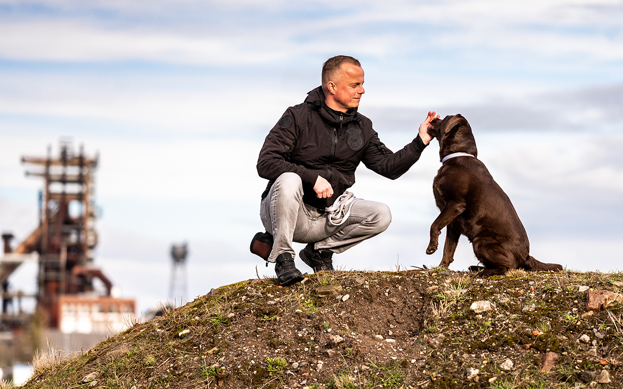 Ein Mann streichelt einen braunen Labrador auf einem Erdhügel. Im Hintergrund ist unscharf ein Industrie-Komplex zu sehen.