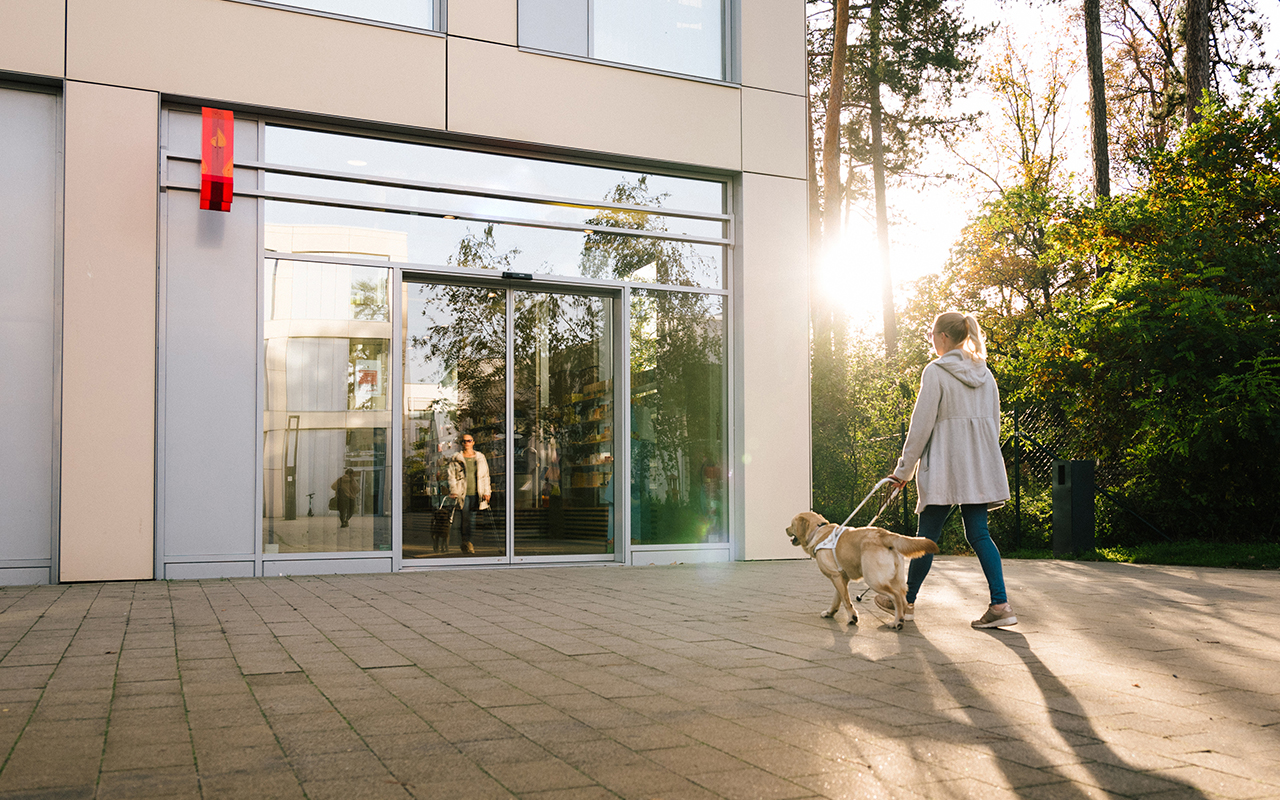 Eine Frau mit Assistenzhund geht auf den Eingang einer Apotheke zu